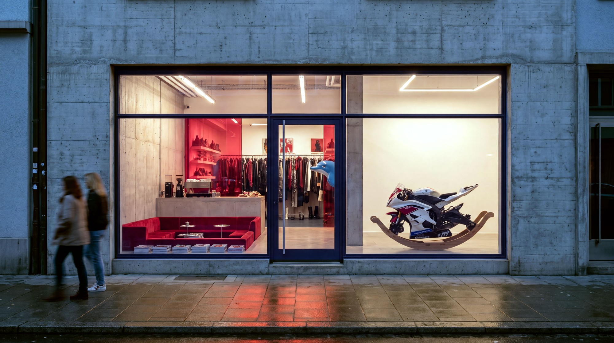 A render of an NMBRD closet on a Zürich street at dusk: a concrete shopfront with a red velvet lounge and a coffee bar, rails of clothes behind the glass door, and a motorbike on rockers in the window.