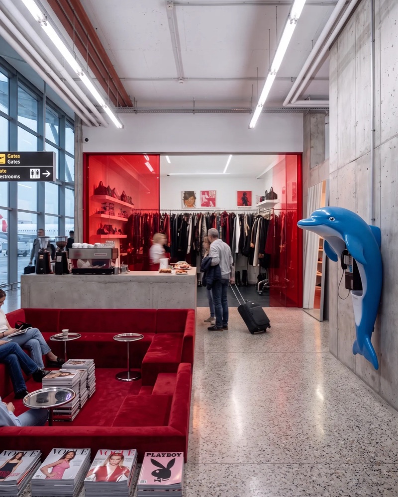 An NMBRD closet at an airport: red glass walls around rails of clothes, a coffee bar, a red velvet seat and a blue dolphin phone on the wall.